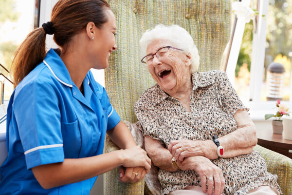 Senior Woman Sitting In Chair And Talking With Nurse In Retirement Home