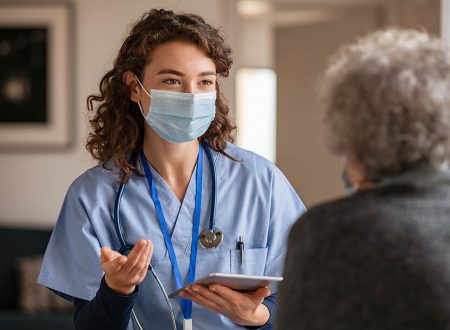 Doctor wearing safety protective mask supporting and cheering up senior patient during home visit during covid-19 pandemic. Nurse and old woman wearing facemasks during coronavirus and flu outbreak.